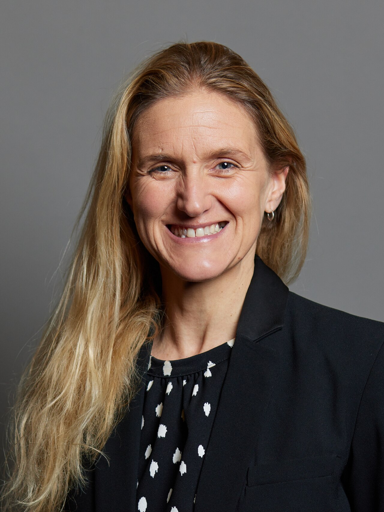 Official portrait of Kim Leadbeater, Member of the UK Parliament, taken from UK Parliament website after her election in 2021. A colour photograph portrait of a white woman with long fair hair, wearing a black jacket over a black and white 